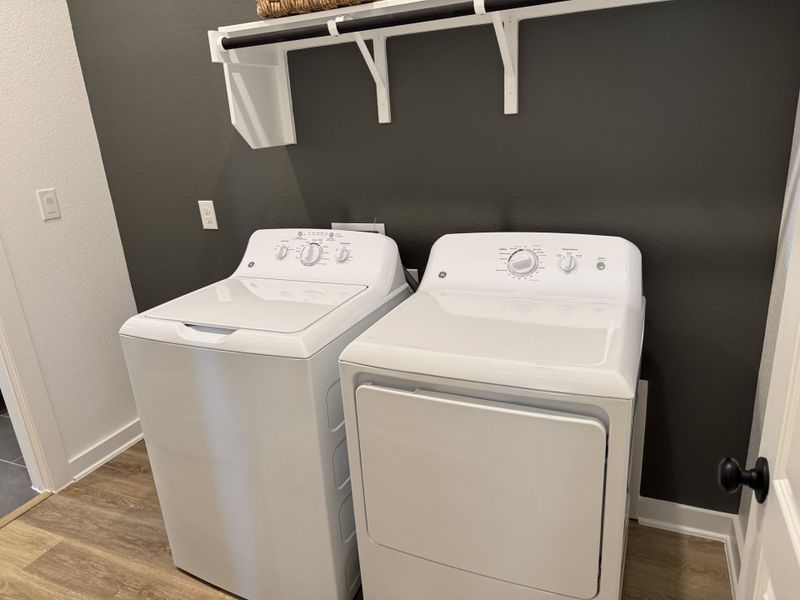 A modern laundry room with sleek white washer and dryer, dark accent wall, and a wooden floor.