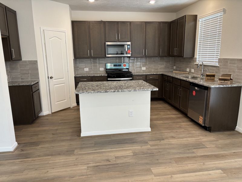 A modern kitchen with dark wood cabinets, granite countertops, and sleek appliances. Light wood flooring complements the space.