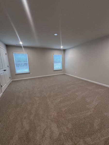 A spacious carpeted room with neutral walls, recessed lighting, and two windows creating a bright and airy atmosphere.