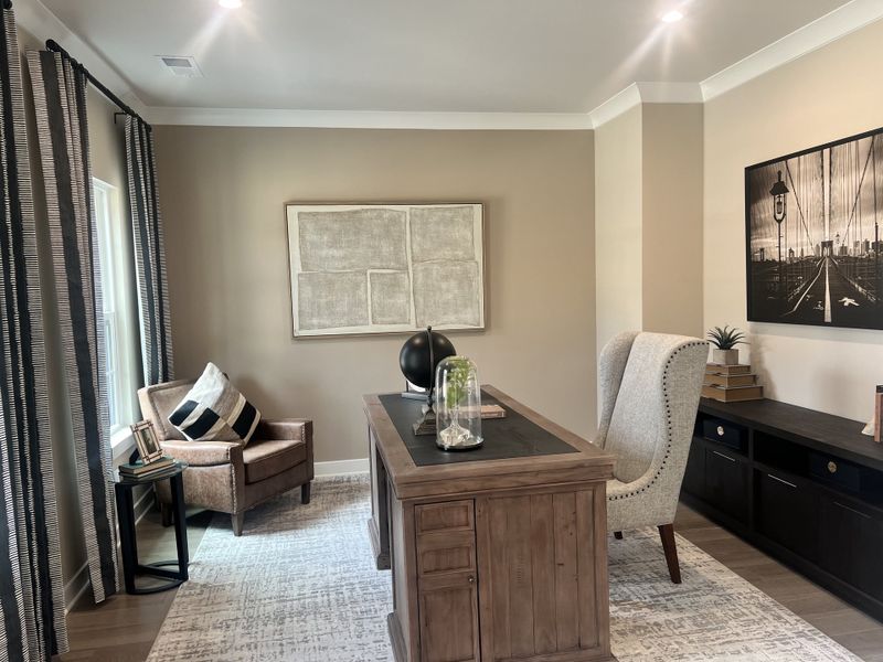 A cozy home office with a wooden desk, plush chairs, modern artwork, and natural lighting through large windows.