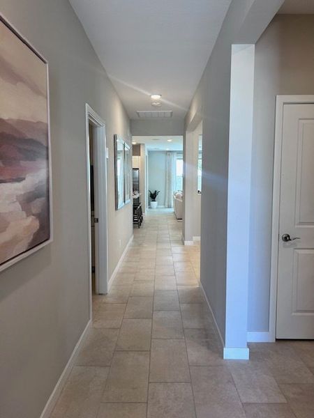 A long, bright hallway with tiled floors, neutral walls, and modern art leading to a sunlit living area.