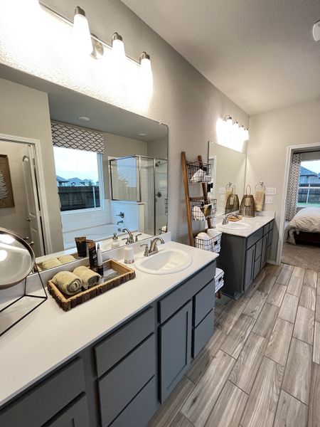 A modern bathroom with dual sinks, sleek gray cabinetry, wood-look tile flooring, and elegant lighting fixtures.