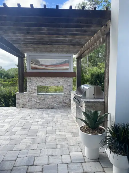A chic outdoor kitchen with a stone fireplace, grill, and modern pergola. Elegant planters add a touch of greenery.