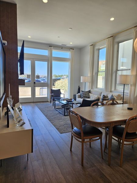 A bright living area with large windows, a cozy seating area, and elegant wooden dining set. Modern and inviting ambiance.