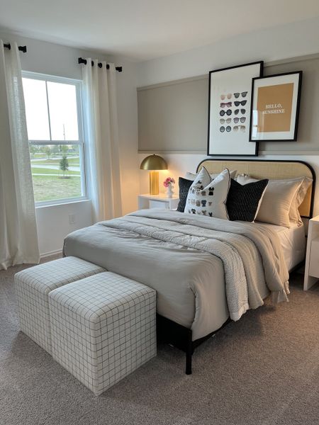 A cozy bedroom with a plush bed, stylish ottomans, gallery wall art, and a warm golden lamp on a nightstand.