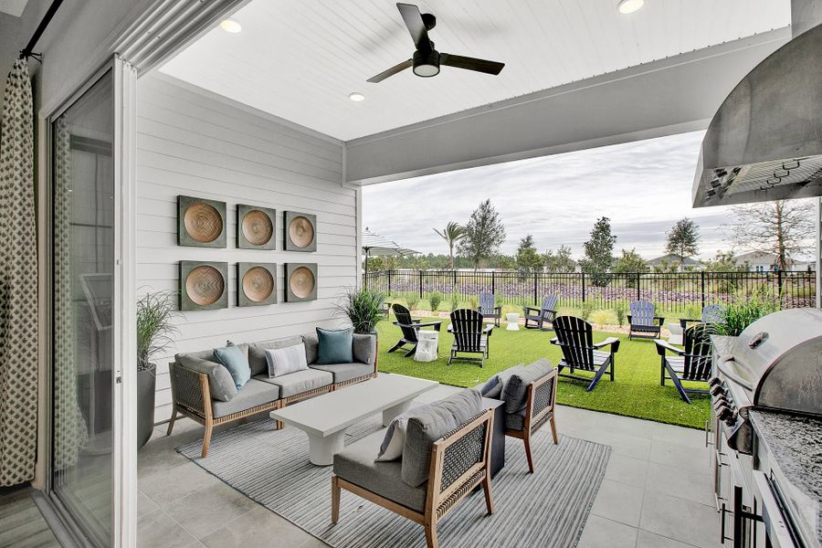 Covered patio with outdoor seating, ceiling fan, built-in grill, and fenced backyard with Adirondack chairs on artificial grass