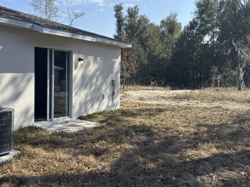 A backyard with a sliding glass door, adjacent to a wooded area, featuring natural landscaping and an air conditioning unit.