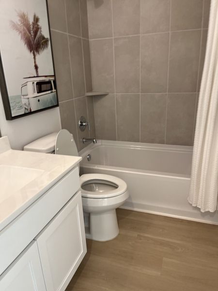 A modern bathroom with gray tiles, white vanity, wood-look flooring, and a coastal-themed wall art. A modern bathroom with gray tiles, white vanity, wood-look flooring, and a coastal-themed wall art.