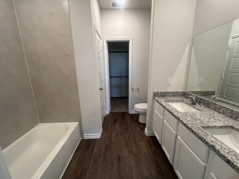 A sleek bathroom featuring a granite-topped double vanity, wood flooring, and a tiled bathtub for modern convenience.