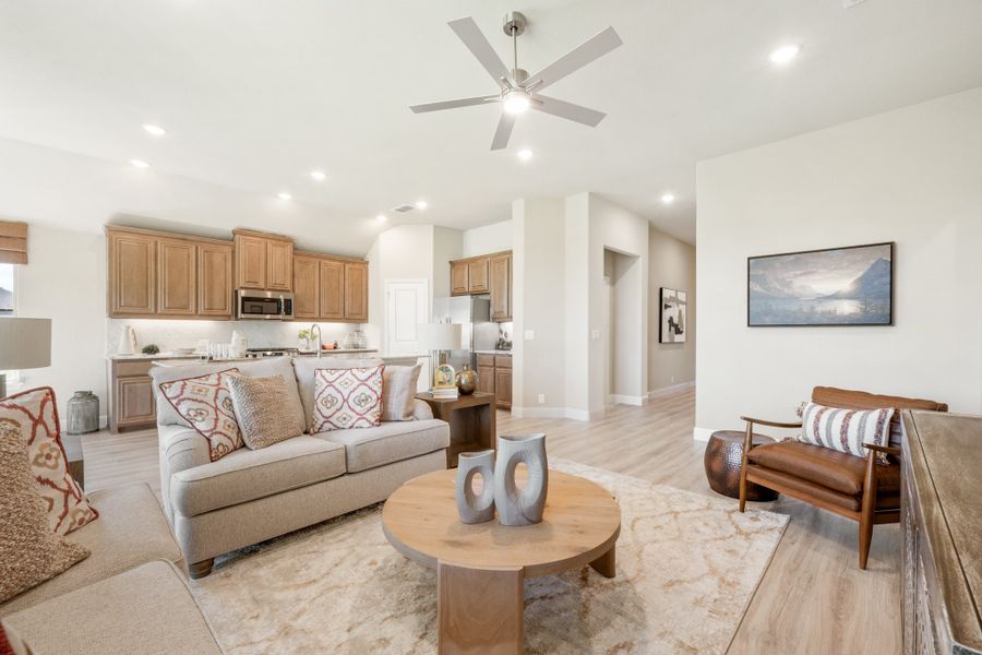 Open-concept living room with ceiling fan and kitchen visible in background, light wood floors and neutral furnishings