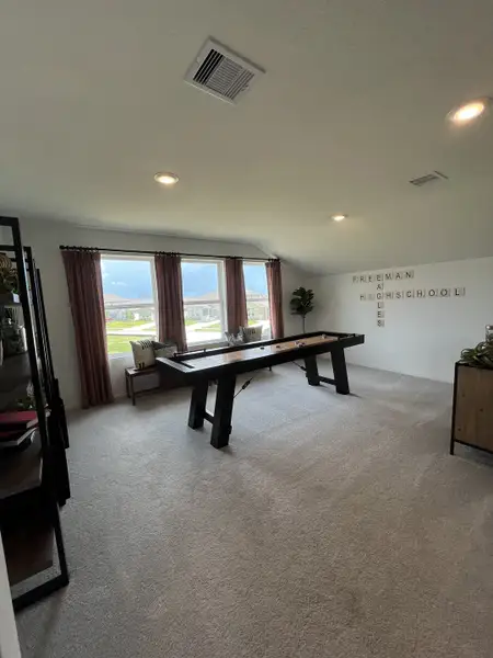 A cozy game room featuring a shuffleboard table, plush carpeting, and ample natural light through large windows.