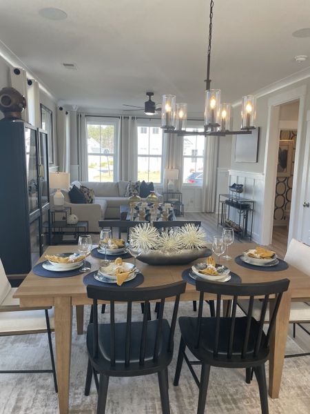 A well-lit dining area with a wooden table set for six, featuring black chairs, elegant place settings, and a view into a comfortable living room with a grey sofa.
