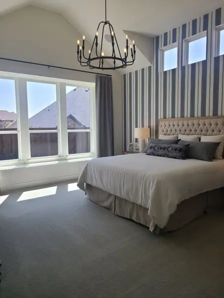 A cozy bedroom featuring a tufted bed, striped accent wall, large windows, and an elegant chandelier.