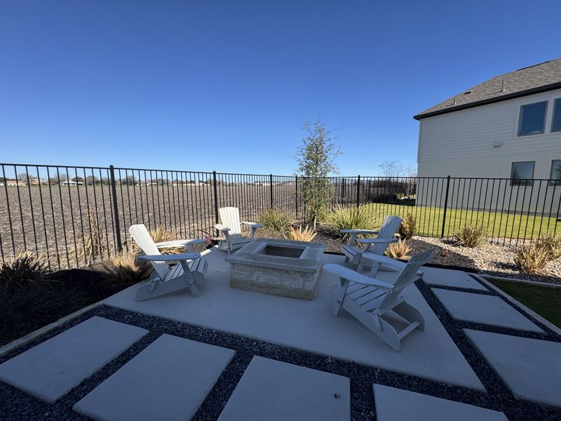 A serene outdoor patio with Adirondack chairs around a stone fire pit, enclosed by a fence overlooking open fields.