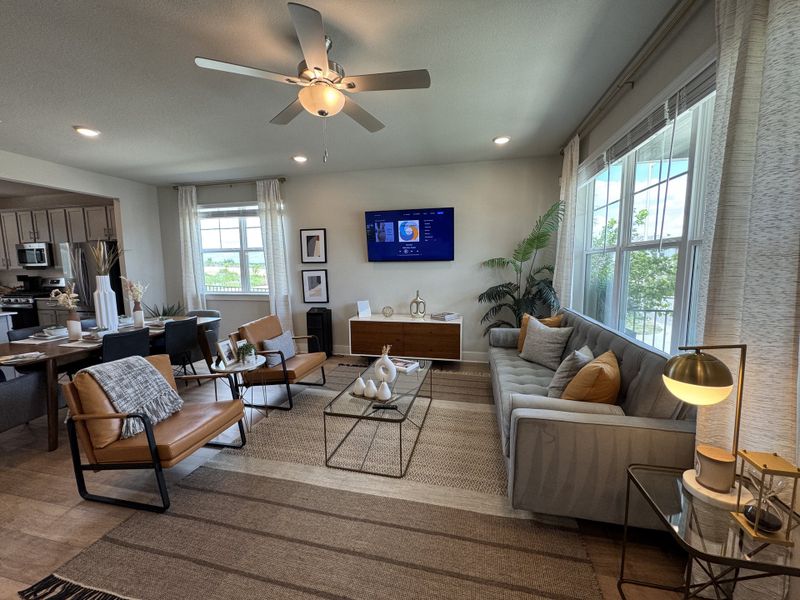A modern living room with a cozy sectional, leather chairs, and a TV. Open layout with dining and kitchen areas visible.