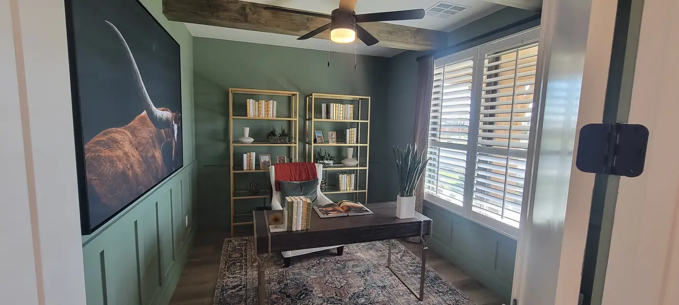 A cozy study with green walls, wooden beams, gold bookshelves, and a plush chair under a ceiling fan with window shutters. A cozy study with green walls, wooden beams, gold bookshelves, and a plush chair under a ceiling fan with window shutters.