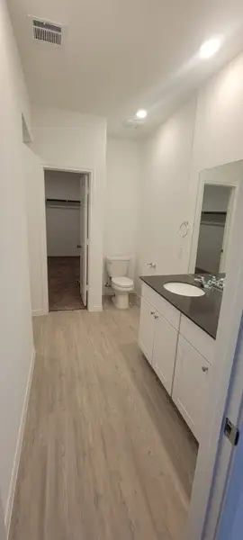A sleek bathroom with light wood flooring, a black countertop, and white cabinets, leading to a walk-in closet.