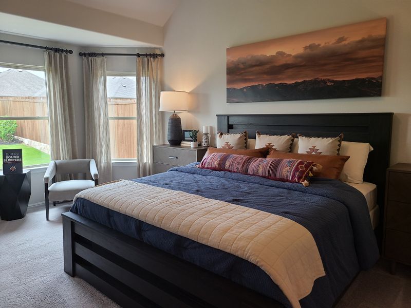 A cozy bedroom with a dark wood bed, mountain artwork, and serene natural light from large windows.