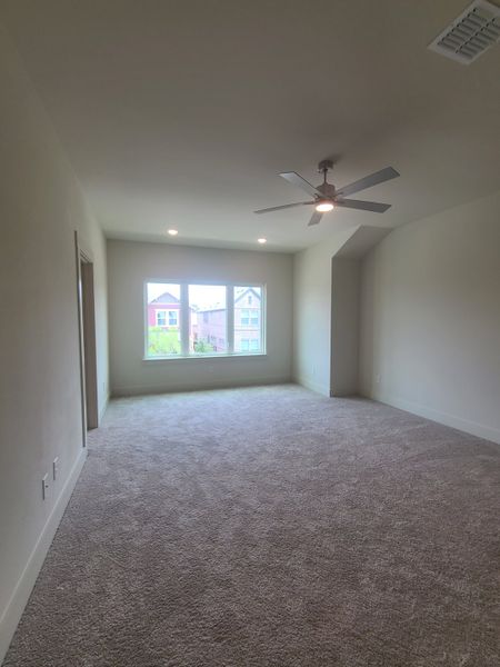 A spacious, carpeted room with neutral walls, a ceiling fan, and large windows for natural light.
