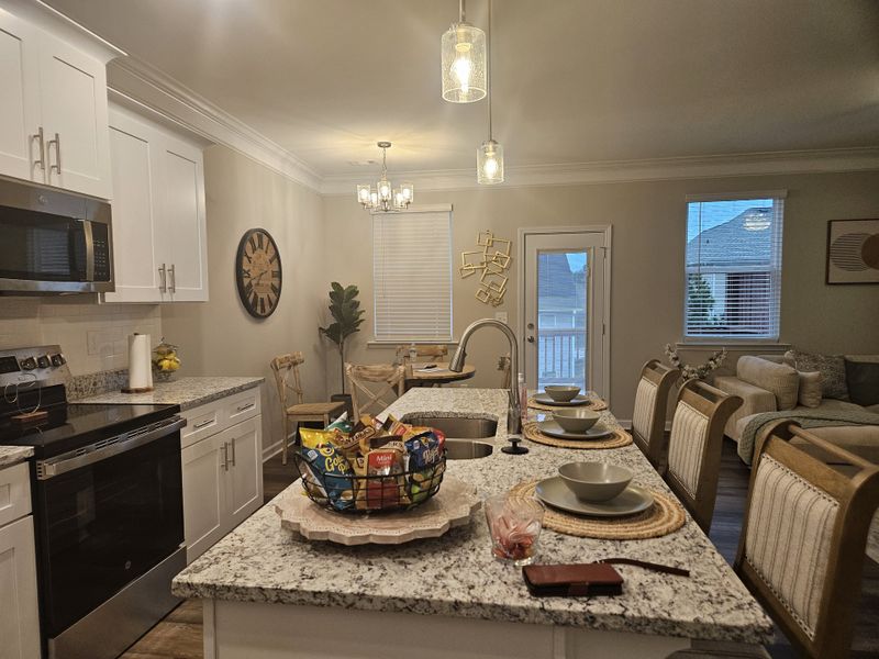 A cozy kitchen with a granite island, set dining table, and pendant lighting, connecting to a living area with neutral decor.