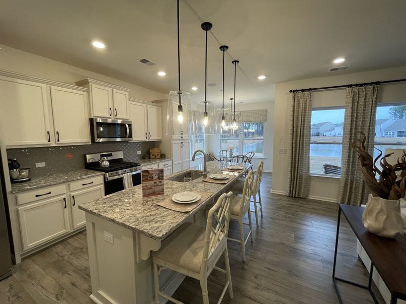 A modern kitchen with granite countertops, elegant lighting, and large windows, featuring white cabinetry and stainless appliances.
