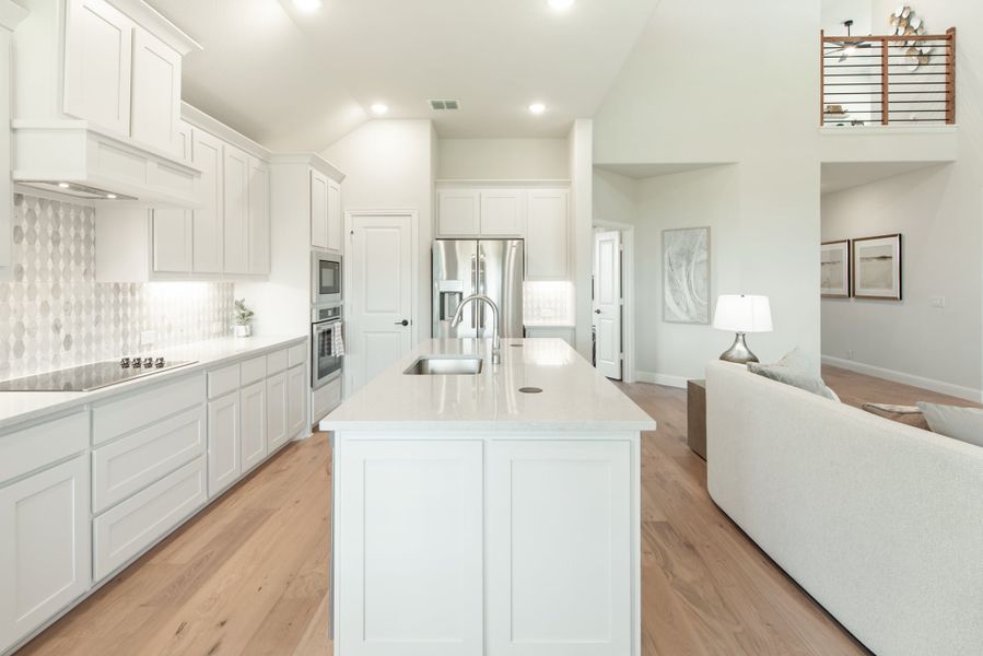 Open kitchen with white cabinets, large island with sink, stainless appliances, and light wood floors opening to living area. Open kitchen with white cabinets, large island with sink, stainless appliances, and light wood floors opening to living area.