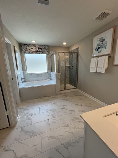 A bright bathroom featuring marble flooring, a glass shower, and a large soaking tub under a soft-lit window.