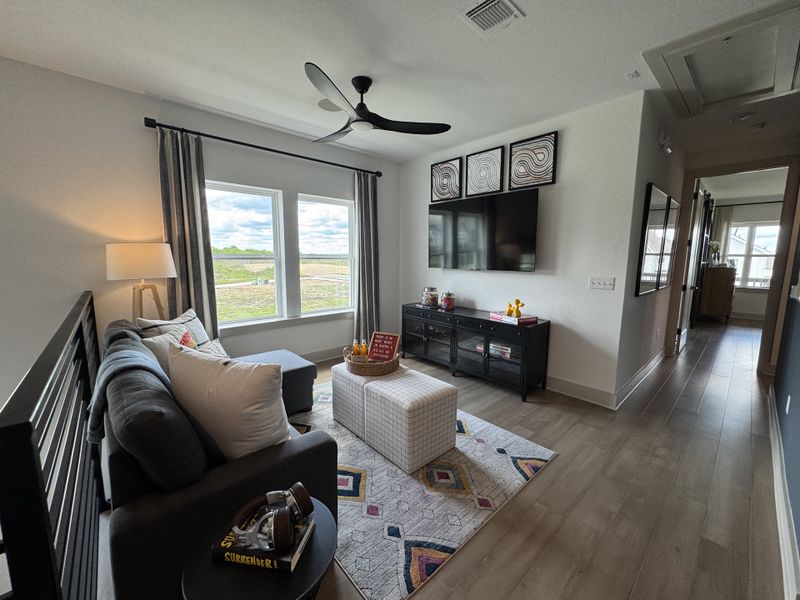 A cozy living area with a gray sofa, large windows, modern fan, and wall-mounted TV, leading to a bright hallway.
