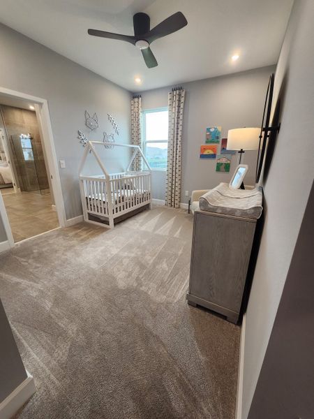 A cozy nursery featuring a sleek ceiling fan, a white house-frame crib, and soft carpeting with modern decor accents.