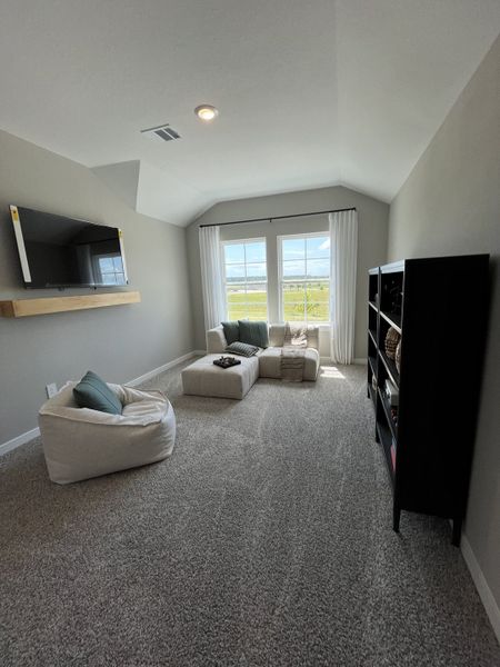A cozy living space with plush carpeting, soft seating, a mounted TV, and natural light from a large window.