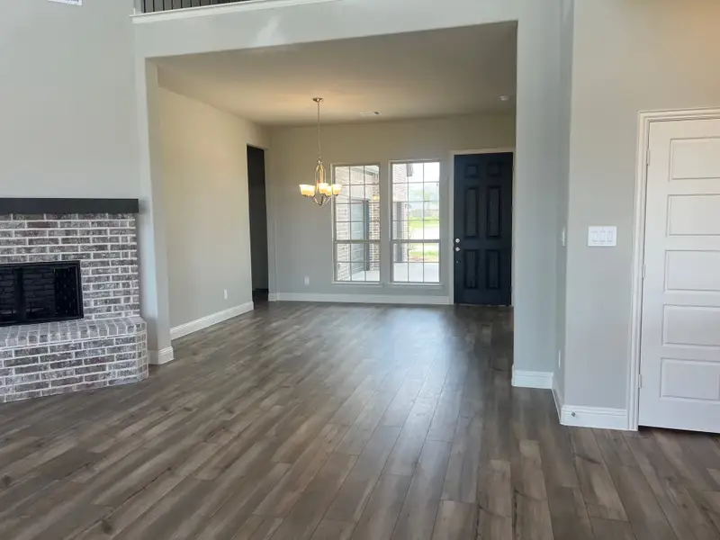 An elegant open living area with wood flooring, a brick fireplace, and ample natural light and overhead lighting.