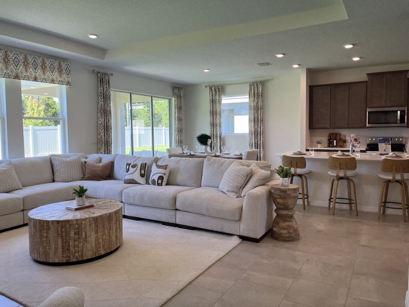 A bright, open-concept living room with a plush sectional sofa, elegant drapes, and a modern kitchen with warm wooden cabinets.