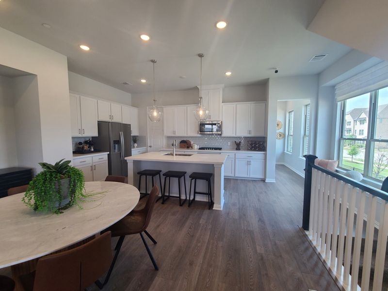 A modern kitchen with white cabinets, island seating, hardwood floors, and sleek lighting, adjacent to a bright dining area.