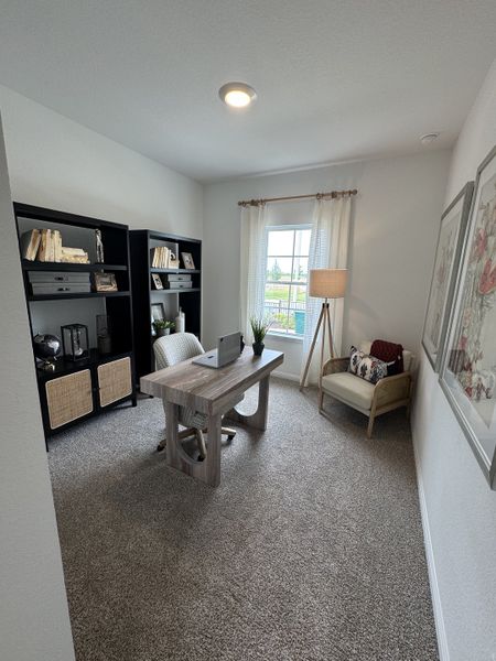 A cozy home office with a modern desk, plush carpet, and ample natural light from a large window.