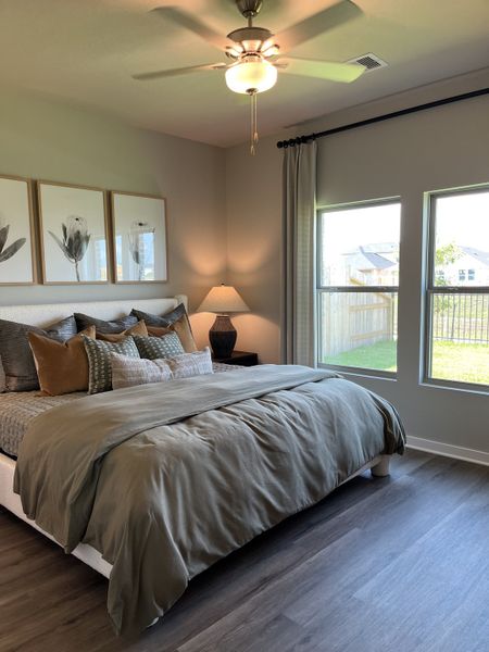 A cozy bedroom with elegant bedding, large windows, modern art, and a ceiling fan, offering ample natural light.