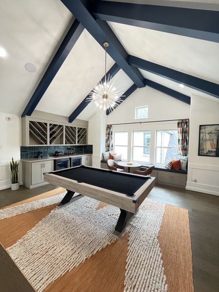 A chic game room with a sleek pool table, vaulted ceilings, bold blue beams, and cozy seating by the window.