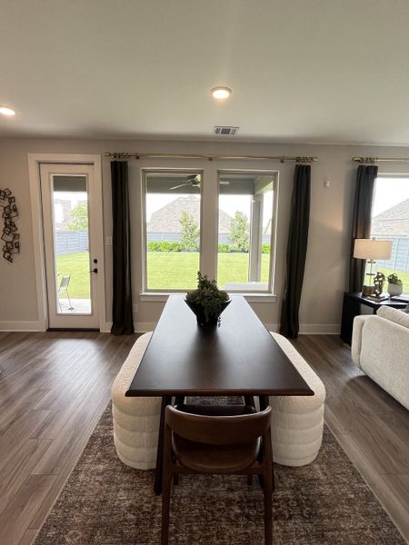 A modern dining area with wood flooring, large windows, and a sleek table. Cozy chairs and greenery complete the look.