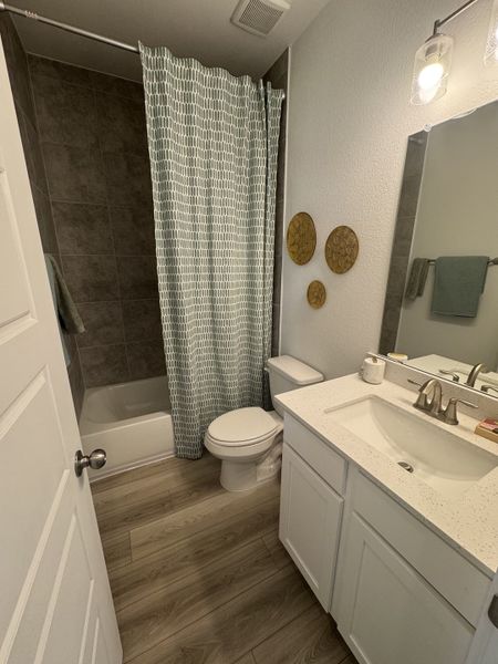 A cozy bathroom featuring a patterned shower curtain, sleek vanity, and warm wood-tone flooring.