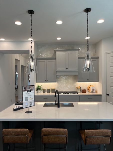 A sleek kitchen with gray cabinets, hexagonal backsplash, pendant lights, and a large white island with bar seating.