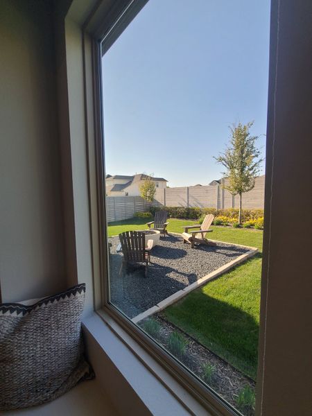 A cozy window view of a tranquil backyard with Adirondack chairs and a fire pit, framed by lush greenery.