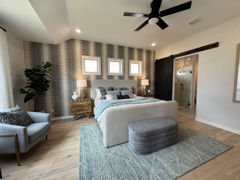 A cozy bedroom with a patterned accent wall, modern ceiling fan, plush seating, and a sliding barn door leading to an ensuite. A cozy bedroom with a patterned accent wall, modern ceiling fan, plush seating, and a sliding barn door leading to an ensuite.