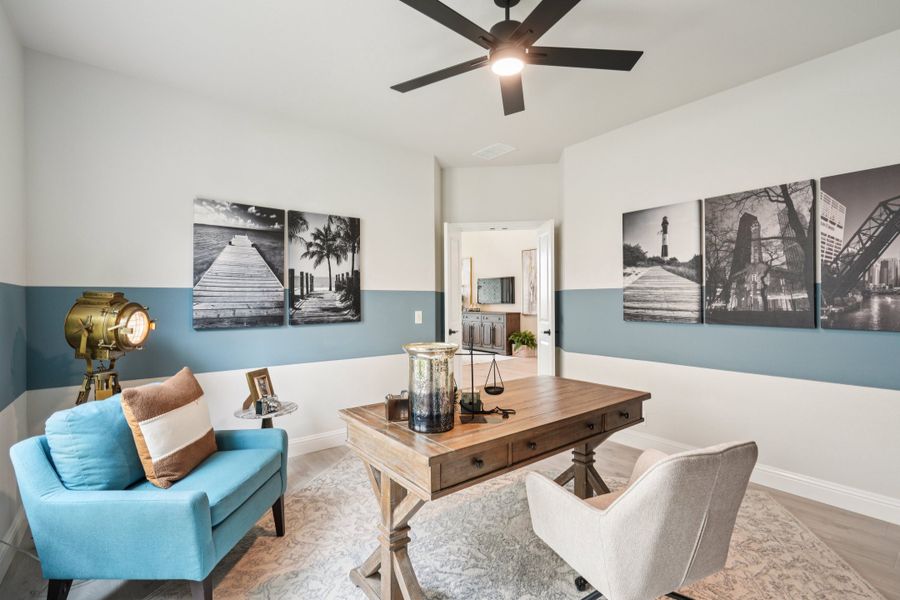 Home office with wooden desk, blue accent walls, ceiling fan, and black-and-white wall art panels Home office with wooden desk, blue accent walls, ceiling fan, and black-and-white wall art panels