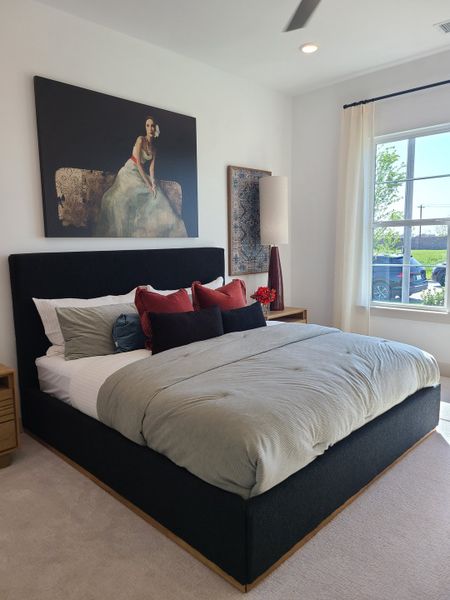 A cozy bedroom with a modern black bed, plush bedding, and elegant artwork above. Bright natural light from a large window.