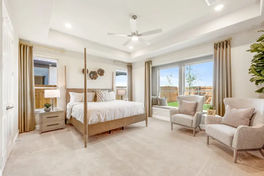 Spacious master bedroom with four-poster bed, tray ceiling, ceiling fan, and window seating area with neutral carpet Spacious master bedroom with four-poster bed, tray ceiling, ceiling fan, and window seating area with neutral carpet