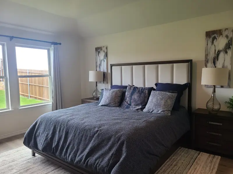 Cozy bedroom featuring a plush headboard, navy bedding, stylish lamps, and large windows overlooking a lush backyard. Cozy bedroom featuring a plush headboard, navy bedding, stylish lamps, and large windows overlooking a lush backyard.