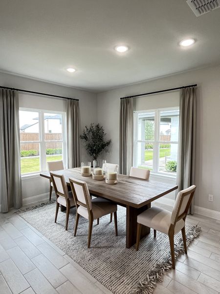 A bright dining room with a wooden table, modern chairs, and large windows. Soft curtains and a textured rug add elegance.