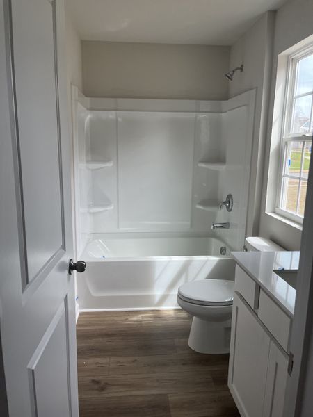 A bright bathroom with a white bathtub, wood-look flooring, and a window providing natural light. A bright bathroom with a white bathtub, wood-look flooring, and a window providing natural light.