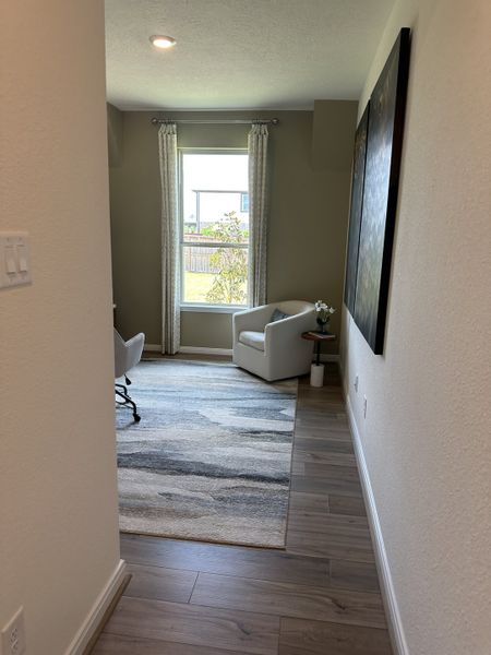 A cozy study nook featuring light wood flooring, an abstract rug, and a modern armchair by a large window with elegant curtains.