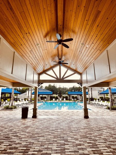 A luxurious pool area with a vaulted wooden ceiling and brick flooring, featuring lounge chairs and blue umbrellas.