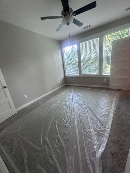 A cozy room with plush carpet, neutral walls, and large windows, complemented by a modern ceiling fan.